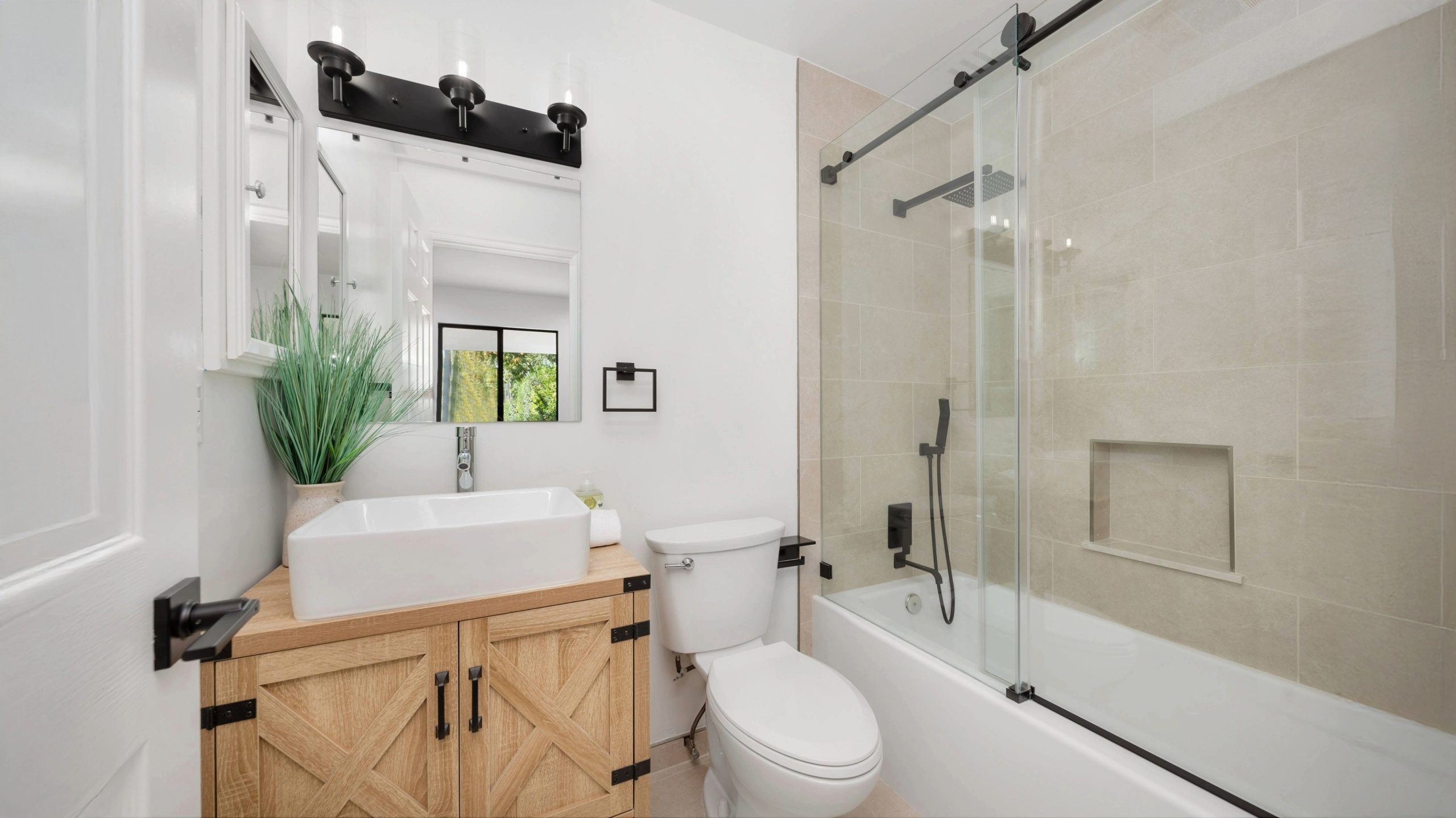 Modern small bathroom remodel featuring a light wood vanity with black hardware, a vessel sink, and a two-piece toilet next to a glass-enclosed bathtub with beige wall tile and matte black fixtures. The space includes a wall mirror, overhead lighting, and a pop of greenery for a clean, contemporary look.