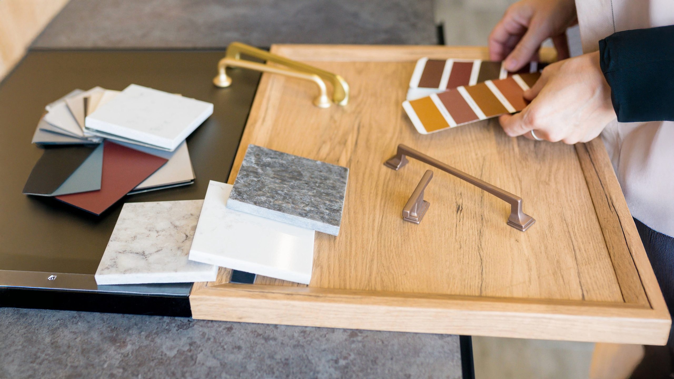 Hands selecting kitchen color swatches and hardware finishes on a wooden tray with countertop samples, paint chips, and cabinet handles during a kitchen remodel cost planning session.