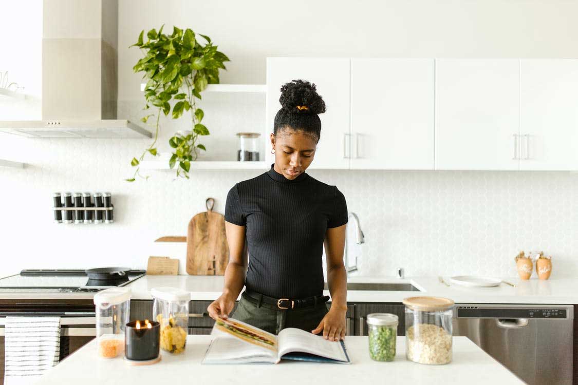 Woman standing in kitchen reading a cookbook on an island with cooking ingredients and scented candle. Leafy green house plant in background.