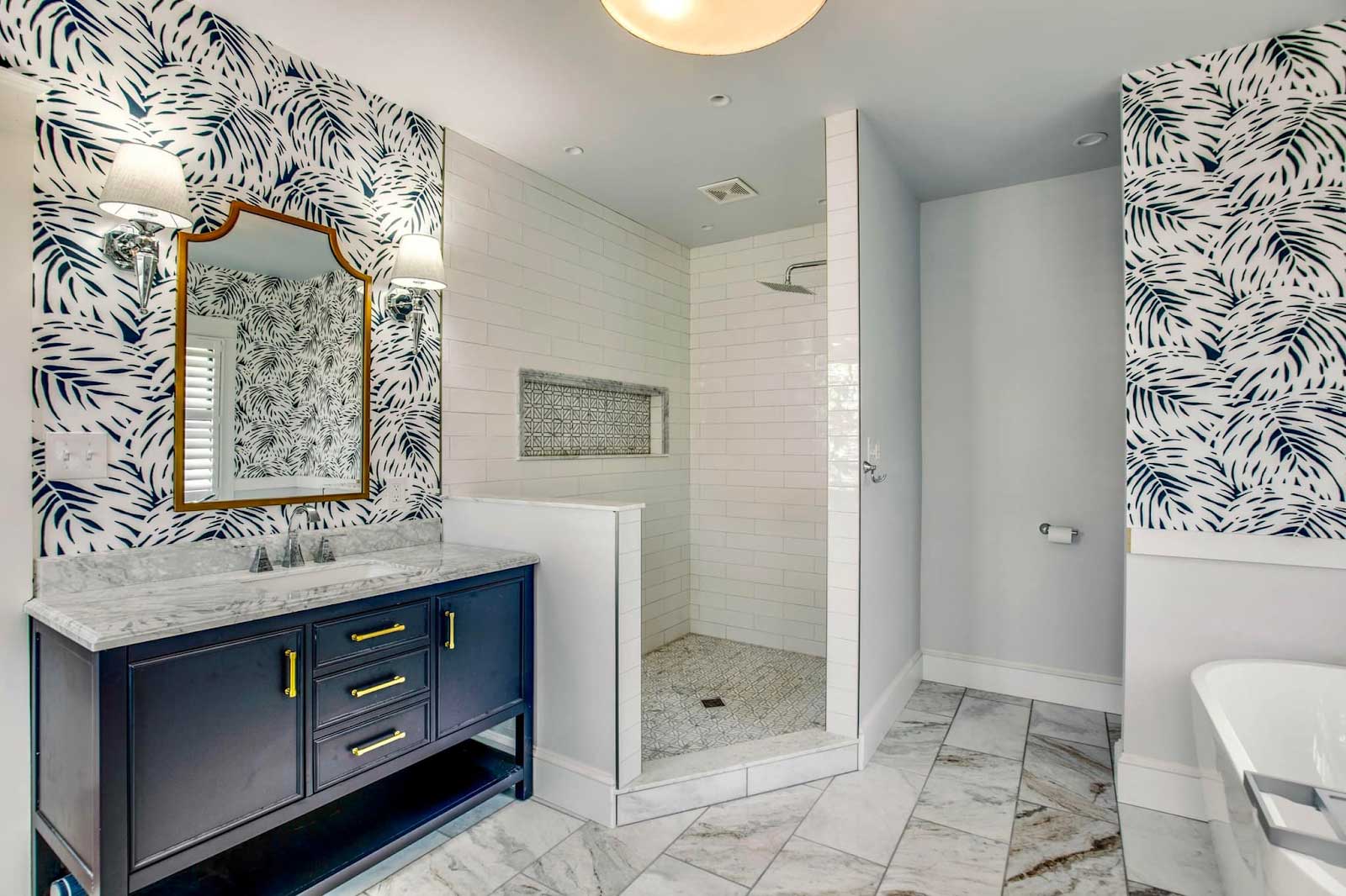 Navy blue bathroom vanity with fun and funky wallpaper next to a huge, tiled shower with niche, water closet, and freestanding bathtub