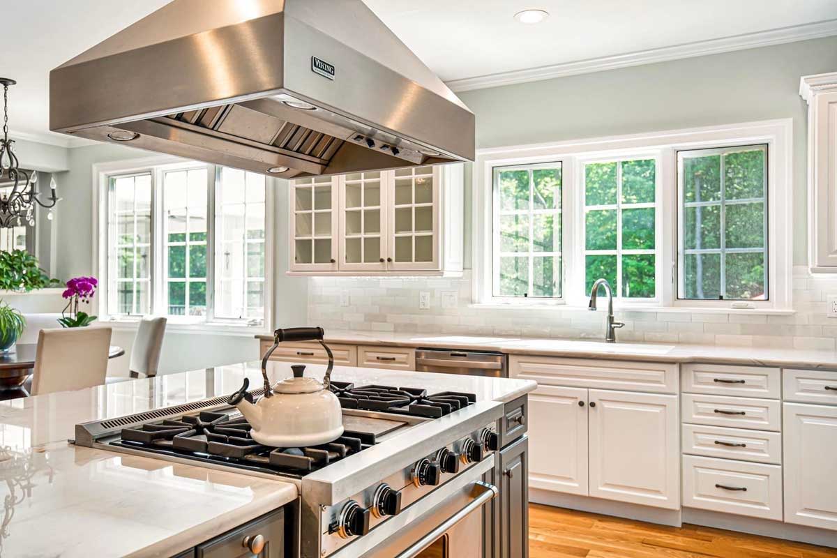 Gas range in kitchen island with overhead exhaust vent and multiple windows over the sink providing natural light and a view