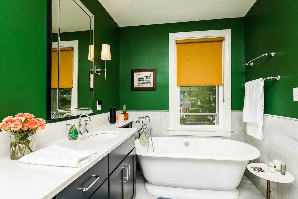 Freestanding bathtub under a window, next to vanity with undermount sink. Bold emerald green paint on the walls add a trendy touch while keeping the fixtures timeless.