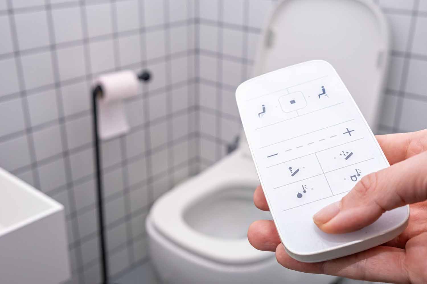 Hand holding remote control with buttons for a smart toilet in the background.