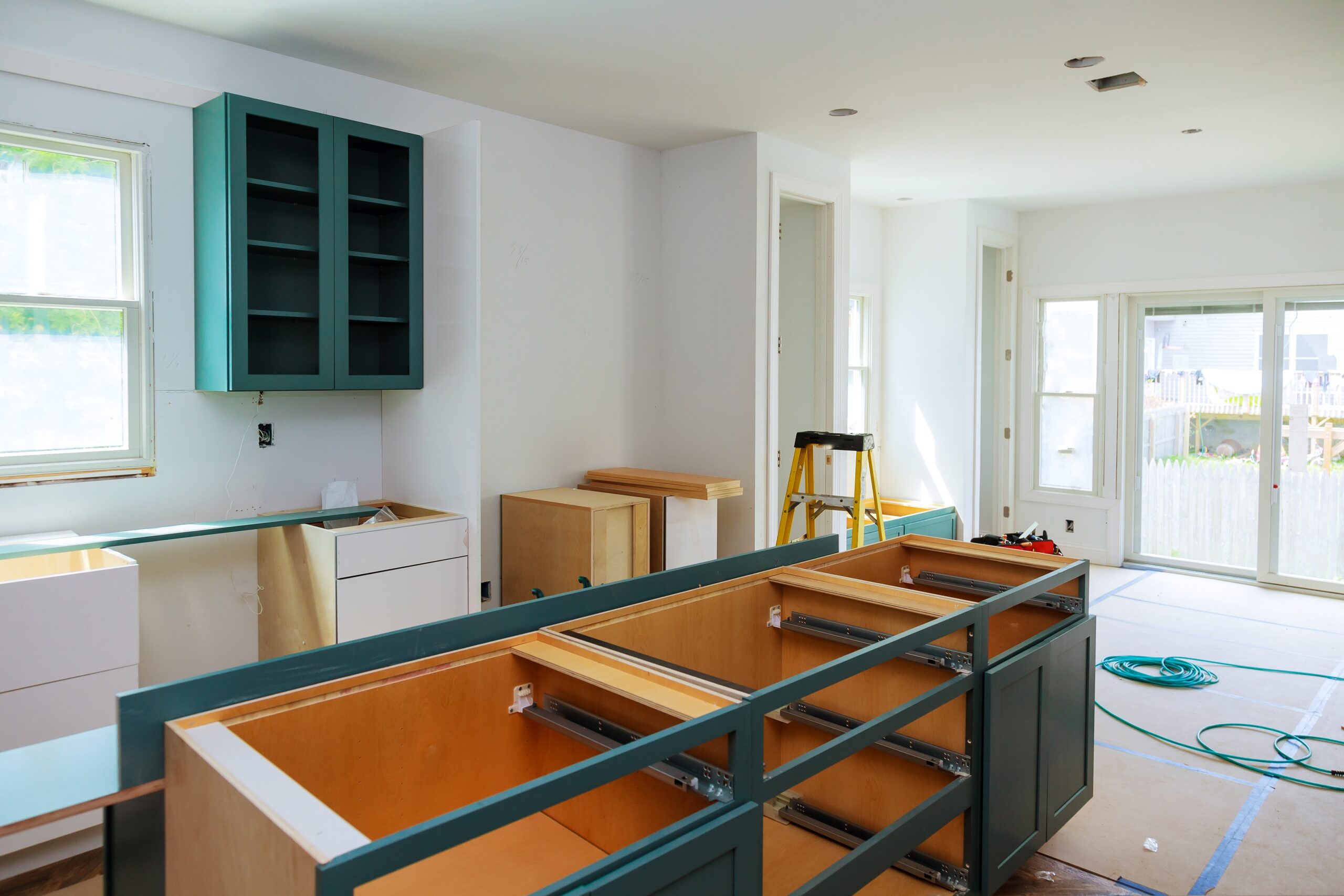 Kitchen under renovation with teal cabinets being installed. A large island without drawers is in the center, and tools, a ladder, and unfinished cabinetry are visible. White walls, large windows, and protective floor covering complete the scene.