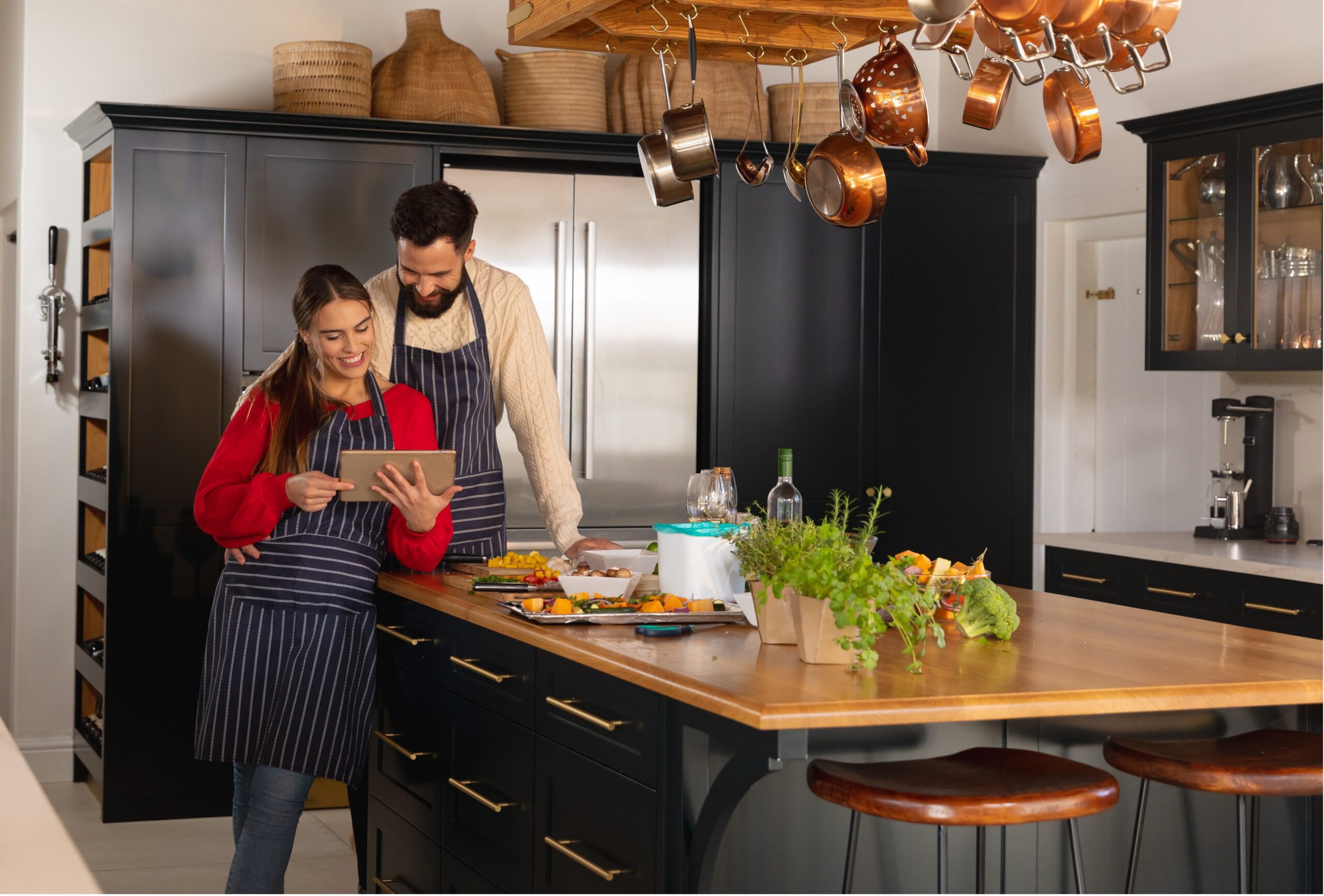 Smiling couple wearing striped aprons stands in a modern kitchen, looking at a tablet while preparing food. The kitchen features dark cabinetry, a wooden island with herbs and vegetables, and hanging copper pots.