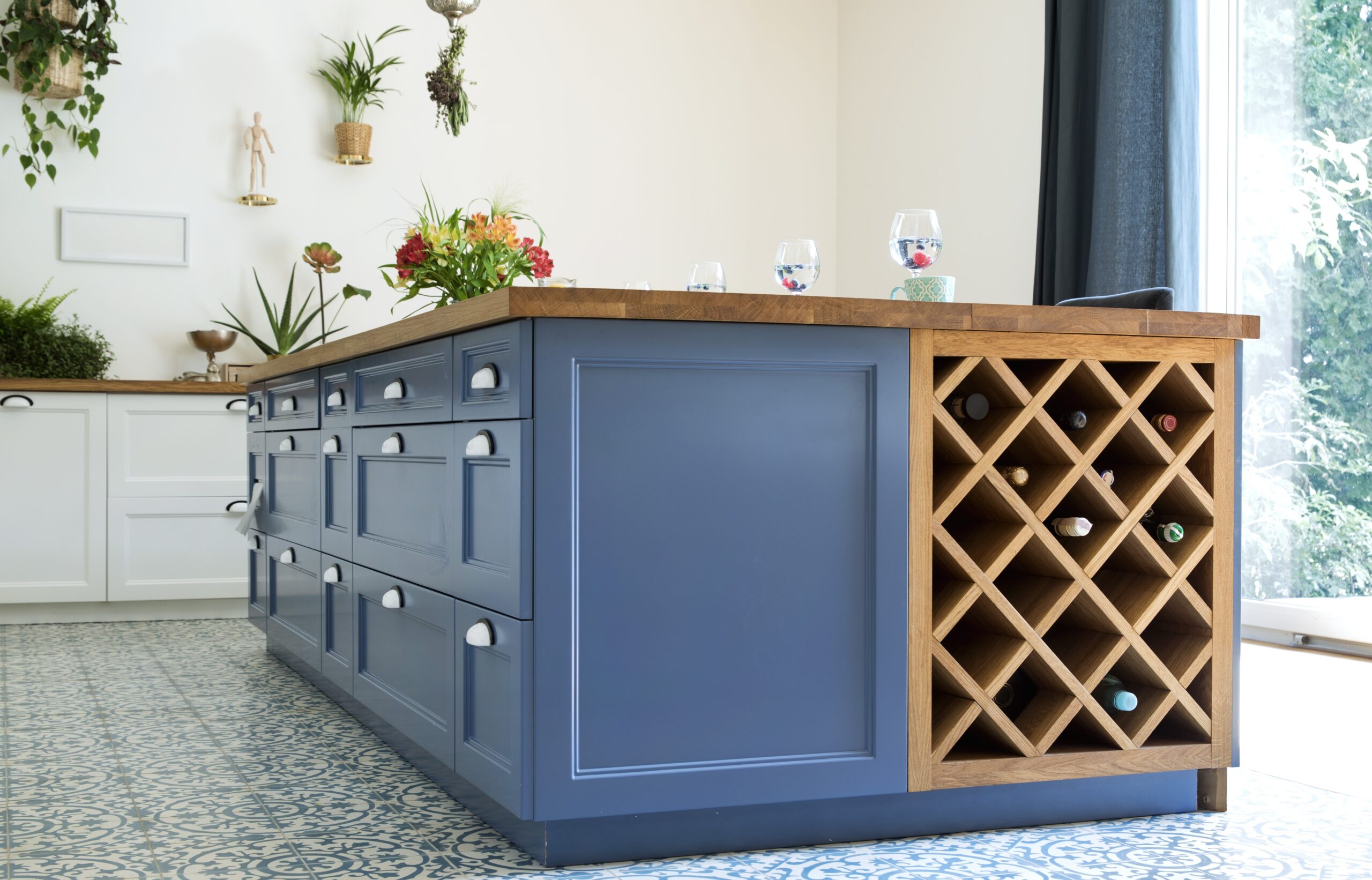 Modern kitchen with a blue island featuring silver cup handles and a built-in wooden wine rack. The island has a butcher block countertop with water glasses and a floral arrangement. The room includes white cabinetry, patterned tile flooring, and hanging plants on the wall.
